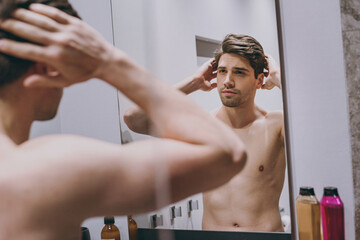 Young smiling handsome happy attractive half naked topless caucasian brunette man 20s look at mirror touch hair indoors in bathroom at home. People healthcare daily morning routine lifestyle concept.