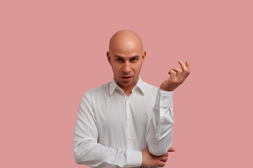 Confused bald guy with bristles hold his hand in question gesture, reacts on strange colleague's phrase, expresses amazed and uncertainty. Dressed in white shirt, isolated over pink ,background. - obrazy, fototapety, plakaty