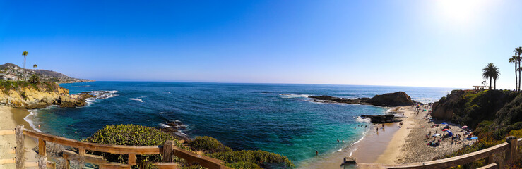 Naklejka na ściany i meble a stunning panoramic shot of the vast blue ocean water with people playing on the sandy beach with lush green hillsides at Treasure Island Beach in Laguna Beach California