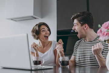 Young couple two woman man fan in casual tshirt clothes sit table drink coffee watch live stream laptop pc computer do winner gesture scream in light kitchen at home together People lifestyle concept