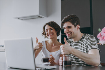 Young couple two woman man in casual t-shirt clothes sit table drink coffee talk zoom video call by laptop pc computer show thumb up gesture in light kitchen at home together People lifestyle concept.