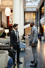 Vertical side view portrait of two successful businessmen discussing work while standing in hotel...