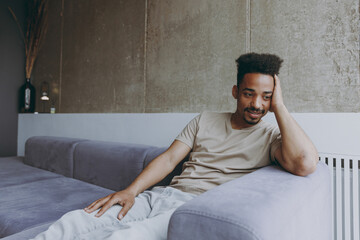 Pensive thoughtful melancholic young african american man in beige t-shirt sweatpants sit on grey sofa indoors apartment near heating battery procrastinate prop up face, rest on weekends stay at home