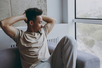 Side view dreamful young african american man 20s in beige t-shirt sweatpants sit on comfortbale grey sofa indoors apartment look at window hold hands behind neck head, rest on weekends stay at home