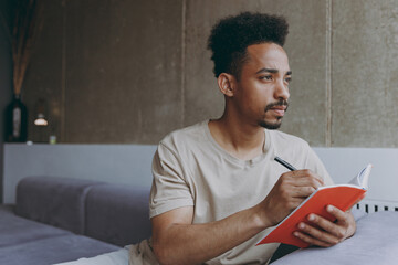 Pensive thoughtful young poet student african american man 20s wearing beige t-shirt sitting on grey sofa indoors apartment write down memories in notebook diary, resting on weekends staying at home - obrazy, fototapety, plakaty
