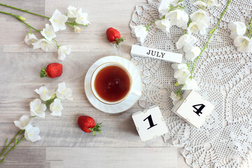 Calendar for July 14 : cubes with the number 14, the name of the month of July in English, a cup of tea on a gray openwork napkin, scattered flowers of bluebells and strawberries, top view