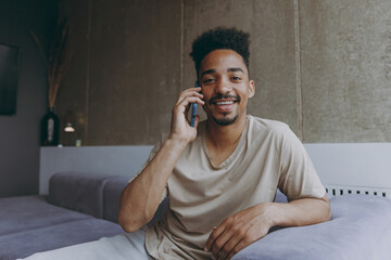 Smiling unshaven young african american man in beige t-shirt sit on grey sofa indoors apartment talk by mobile cell phone with friends look camera rest on weekends stay at home Tattoo translate fun.