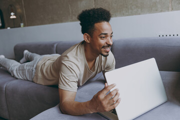 Side view young impressed fun african american man wearing beige t-shirt sweatpants lay down on grey sofa indoors apartment use laptop pc computer look aside resting on quarantine weekends stay home.