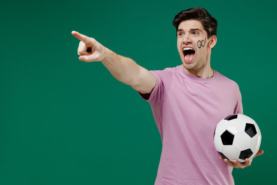 Young fun european man fan supporter wears basic pink t-shirt cheer up support football sport team hold in hand soccer ball watch tv live stream scream isolated on dark green color background studio.