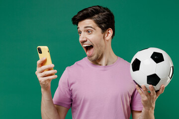 Young fun man fan wear basic pink t-shirt cheer up support football sport team hold in hand mobile cell phone soccer ball watch tv live stream scream isolated on dark green color background studio © ViDi Studio