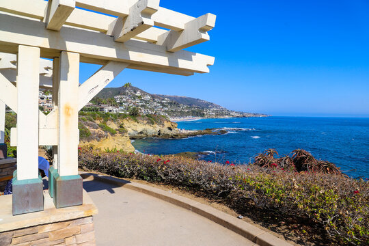a wooden white pergola surrounded by lush green palm trees and plants with red flowers and lush green grass near blue ocean water and blue sky at Treasure Island Beach in Laguna Beach California