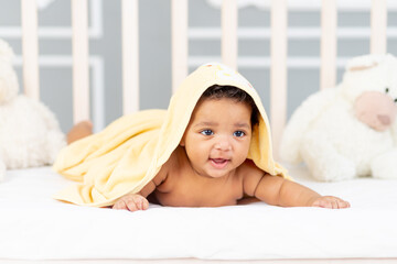 Smiling African-American little baby lies in bed after a bath in a yellow towel