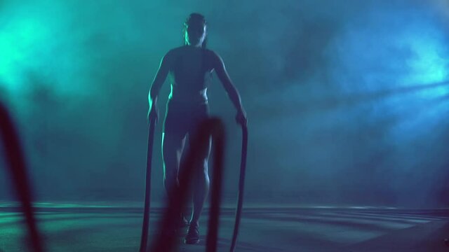 Front view of muscular brunette woman training in gym with ropes in dark atmosphere and smoke. Pretty fitnesswoman with strong face doing power cardio and building muscles. Concept of sport.