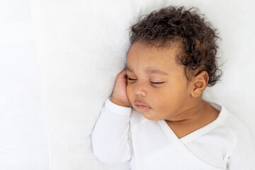 African-American little baby sleeps on a white bed at home with her hand folded under her cheek