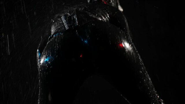 A woman dressed in a tight black latex suit for BDSM role playing games poses and dances close up from the back on a black background on a jet of water