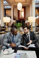 Vertical portrait of two successful businessmen discussing deal while sitting at table in hotel...