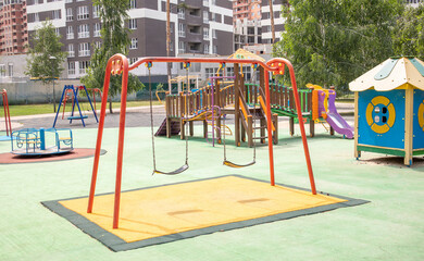 A colorful modern playground in the courtyard in a park without people. Swing, outdoor carousel for children. A playground in a public park surrounded by green trees and new houses. Children exercise.