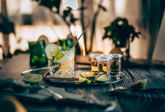 Dark photography of a cold drink, a glass of mojito with mint leaf, ice, lemon and sugar with tequila shots on a wooden table