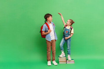 happy teenage schoolboy with a backpack, measuring the height of a little girl in loose clothes, standing on a stack of books on a green background. The concept of development and growth of education. - obrazy, fototapety, plakaty