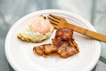 Macro closeup of cooked bacon strips and fried egg on white plate and wooden fork at breakfast brunch traditional American meal