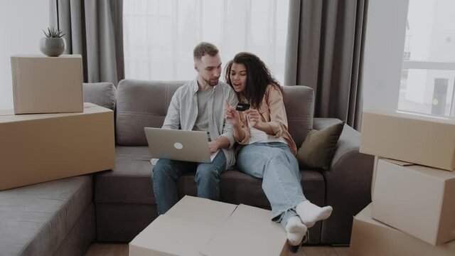 Happy couple beween a heap of boxes in living room sitting at the couch use a laptop and credit card for buying on-line, choose furniture on webstore, search design interior ideas fot their new home