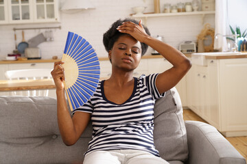 Tired African American woman suffering from heatstroke flat without air-conditioner, waving blue...