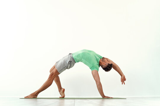 Fit male practicing yoga Wild Thing Pose, Camatkarasana against white wall background