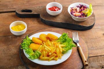 French fries with chicken nuggets next to cheese sauce and ketchup in a gravy boat and salad on wooden planks next to a fork.