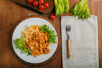 A plate of bolognese pasta decorated with greens on a wooden table next to a gray napkin and a fork and cherry tomatoes.