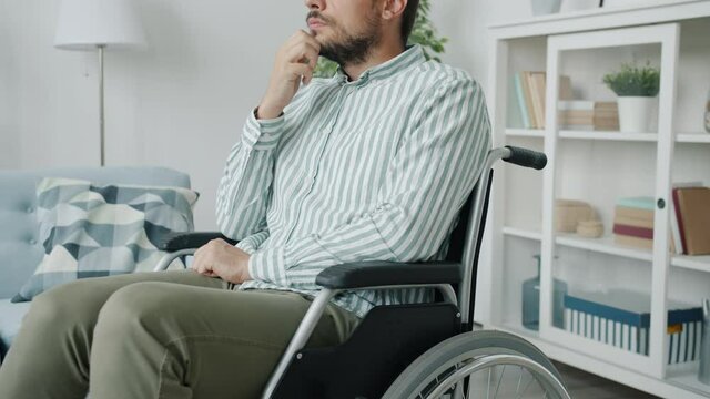 Slow motion portrait of handicapped man sitting in wheelchair in apartment thinking touching beard feeling sad and lonely. People and lifestyle concept.