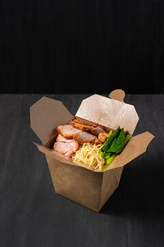 Noodle with pork, meat, vegetable in cardboard box for takeaway with chopstick in dark or black background, asian traditional chinese cuisine food for delivery concept with copy space