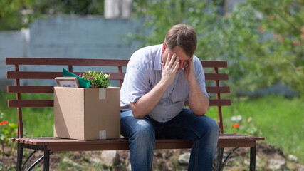 Fototapeta premium A man fired from his job sits frustrated on a bench in a park holding his head.