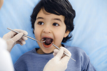 Cute arab boy sitting at dental chair with open mouth during oral checking up with doctor. Visiting...