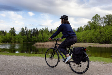 Fototapeta premium Adult Caucasian Woman Riding a Bicycle on a path by a lake in a modern city park. Spring Evening. Taken in Green Timbers Urban Forest, Surrey, Vancouver, British Columbia, Canada.