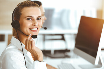 Blond female customer service representative is consulting clients online using headset in sunny...