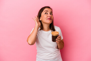 Middle age caucasian woman holding a coffee jar isolated on pink background trying to listening a gossip.