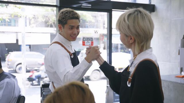 Cheerful young Asian man and woman hairdresser wearing apron uniform in modern salon giving high-five to each other on celebrating success of salon