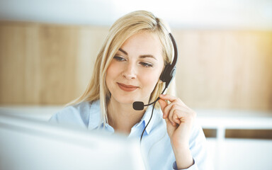 Naklejka premium Blonde business woman sitting and communicated by headset in call center in sunny office. Concept of telesales business