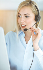 Blonde business woman sitting and communicated by headset in call center in sunny office. Concept...