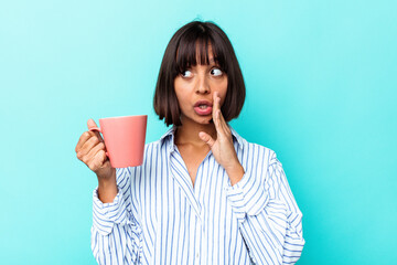 Young mixed race woman holding a pink mug isolated on blue background is saying a secret hot...