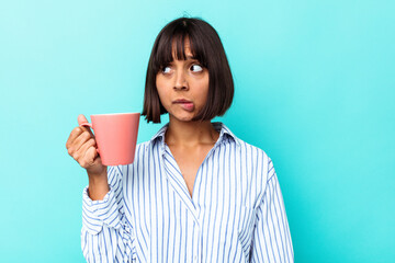 Young mixed race woman holding a pink mug isolated on blue background confused, feels doubtful and unsure.
