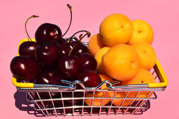 Fresh cherries and apricots on a pink background in a supermarket grocery basket with yellow handles. Selective focus.