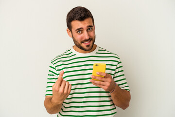 Young caucasian man holding a mobile phone isolated on white background pointing with finger at you as if inviting come closer.