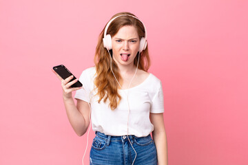 pretty red head woman feeling disgusted and irritated and tongue out with headphones and smartphone