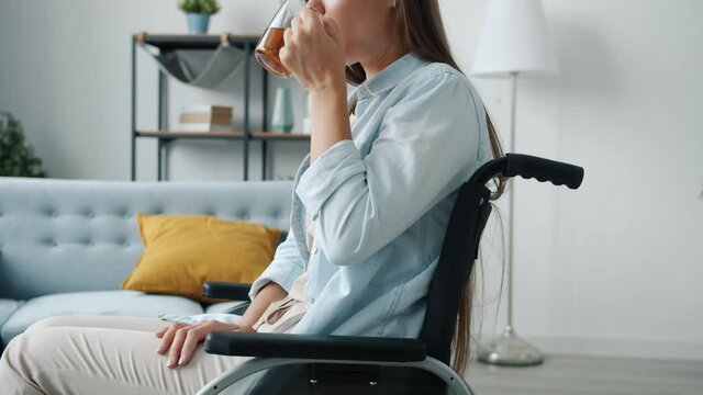 Slow motion portrait of young disabled lady drinking tea sitting in wheelchair alone in apartment. Physical disability and leisure time concept.
