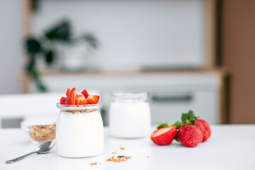 Greek yogurt in a glass jar with muesli and strawberries at the white table.