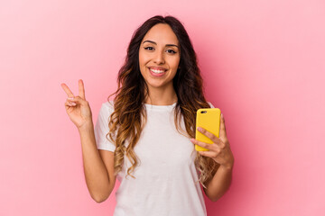 Young mexican woman holding a mobile phone isolated on pink background joyful and carefree showing a peace symbol with fingers.