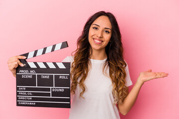 Young mexican woman holding clapperboard isolated on pink background showing a copy space on a palm and holding another hand on waist.