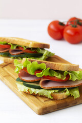 two toasted homemade sandwiches with fresh vegetables on the cutting board. tasty sandwich with ham, green salad, cucumbers and tomatoes isolated on the white background. copy space. vertical