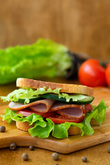 close up tasty sandwich with ham, green salad, cucumbers and tomatoes on the wooden background.toasted homemade sandwich with fresh ingredients on the brown cutting board.vertical. copy space